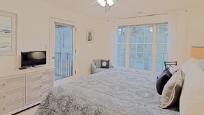 A bedroom with a bed, a TV, a ceiling fan, and large windows.