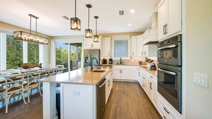 Une cuisine moderne dotée d’un grand îlot central, de meubles blancs, d’appareils électroménagers en acier inoxydable et de luminaires suspendus.