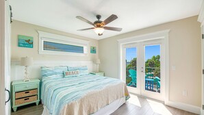 Une chambre à coucher comprenant un lit, deux tables de chevet, un ventilateur de plafond, une fenêtre avec des stores et une vue sur l’extérieur.