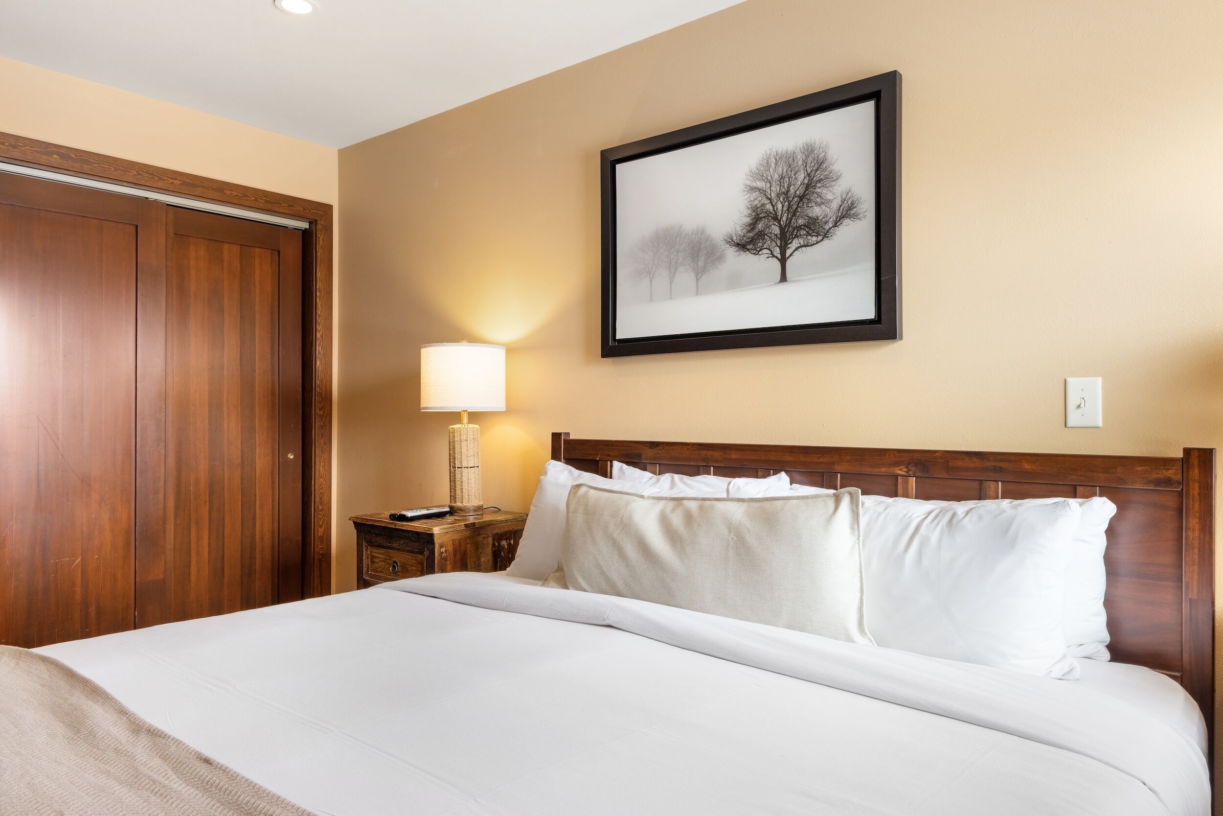 Quarto de hotel com cama de madeira, mesa de cabeceira com abajur e quadro na parede.