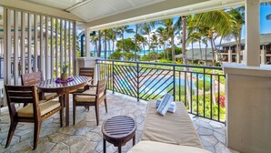 A balcony with a pool, wooden furniture, and a view of the ocean and palm trees.