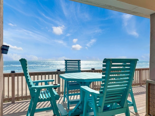 Une terrasse en bois avec des chaises turquoise et une table, donnant sur une plage, l’océan et un ciel dégagé.
