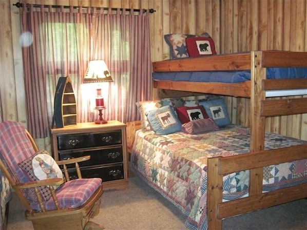 A cozy room with a bunk bed, a chair with a plaid cushion, a dresser with drawers, and a lamp on a wooden nightstand.