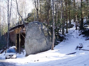A cabin with a large rock in front, surrounded by trees and snow.