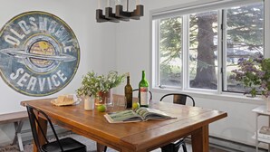Un comedor con una mesa de madera, sillas, un letrero vintage grande, una ventana con vista a un árbol y una lámpara colgante.