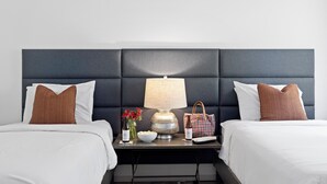 Habitación de hotel con cabecera oscura, dos camas con sábanas blancas, una mesita de noche con lámpara, un cuenco de aperitivos, una botella de vino, un florero con flores y un bolso a cuadros.