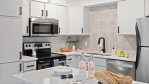 Una cocina moderna con armarios blancos, electrodomésticos de acero inoxidable, una isla central con platos, vasos y botellas, y una lámpara colgante.