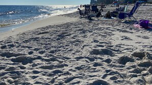 Ein sandiger Strand mit klarem Himmel und strahlender Sonne. Im Hintergrund sind Gebäude zu sehen, sowie Strandstühle und Sonnenschirme.