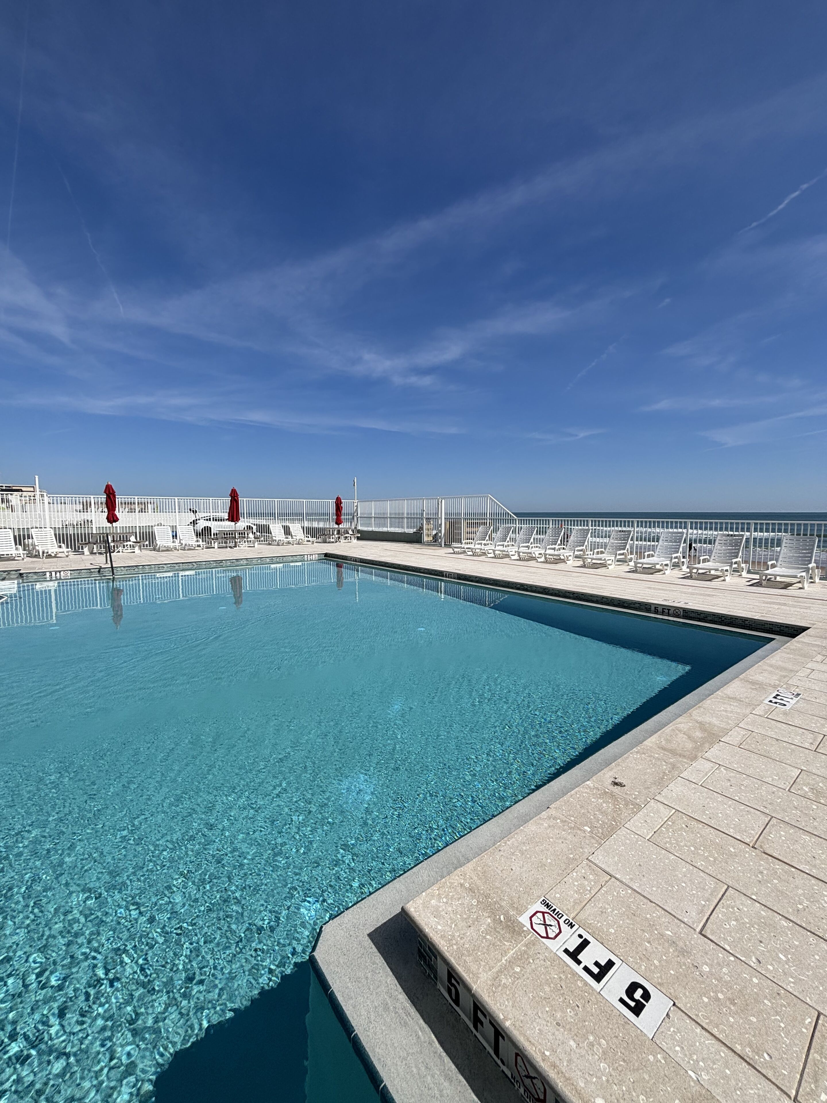 Une piscine d’un bleu éclatant, bordée d’une terrasse blanche et dotée de chaises longues, le tout entouré d’une clôture blanche.