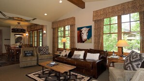 A cozy living room with a brown leather sofa, a wooden coffee table, and patterned cushions. There is a painting of a cow on the wall, and the room has large windows with curtains. - Perfect location makes for easy access to downtown, Snow King and more (Jackson)