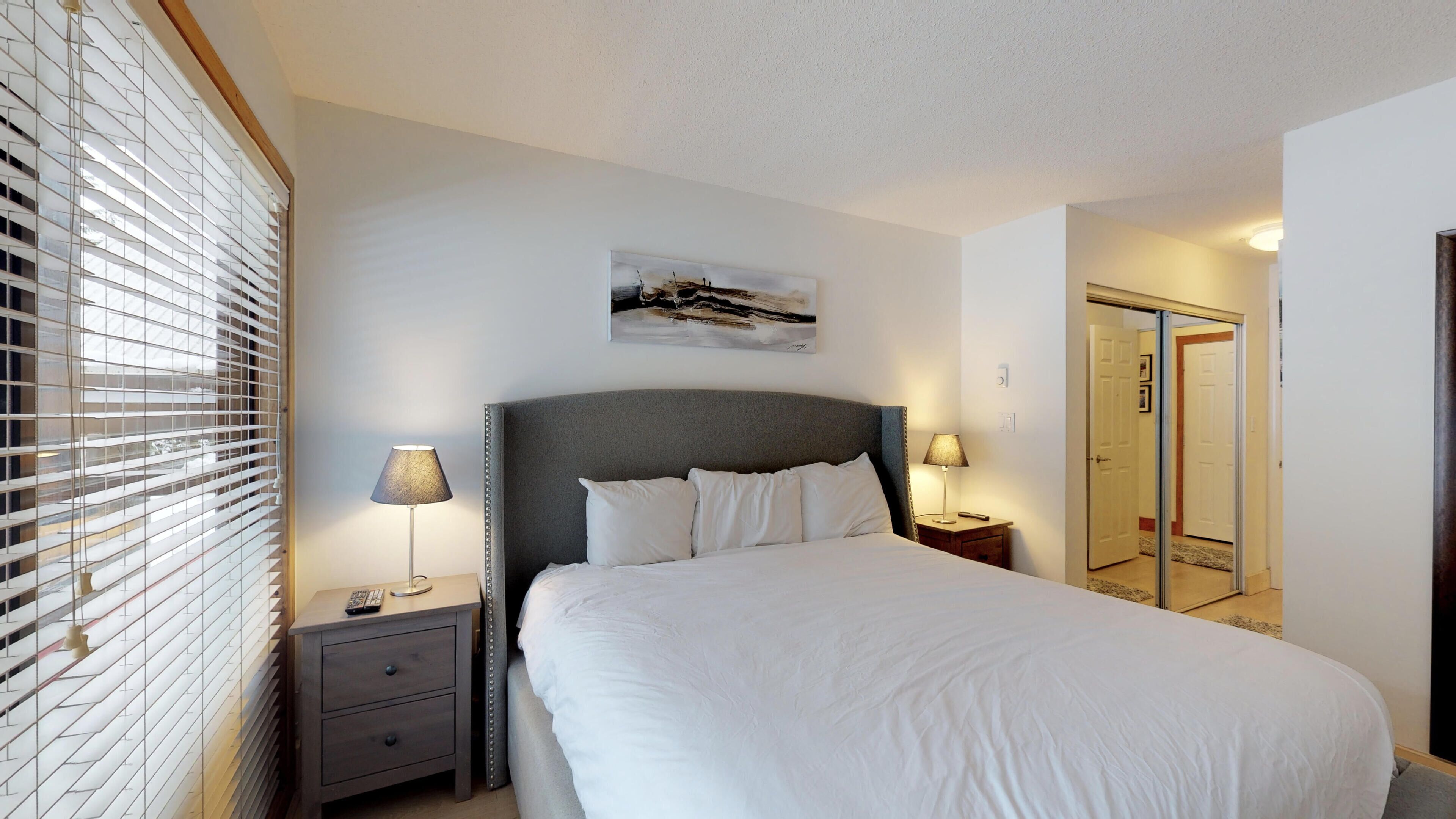 Een netjes ingerichte hotelkamer met een groot bed, bedlampjes, een nachtkastje, een schilderij aan de muur en een deur naar een andere kamer.