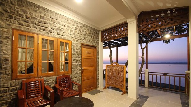 Quarto com paredes de pedra, porta de madeira, duas cadeiras de madeira, uma pequena mesa e vista para o mar ao entardecer.