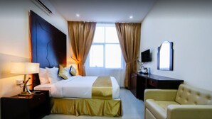 A hotel room with a bed, a bedside table with a lamp, a TV, a sofa, and a window with curtains. - Sh Hotel (Ras Al Khaimah)