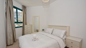 A neatly made bed with rolled towels, a bedside table, a nightstand, a mirror, and a window with curtains. - The Kings Towers (Tiberias)
