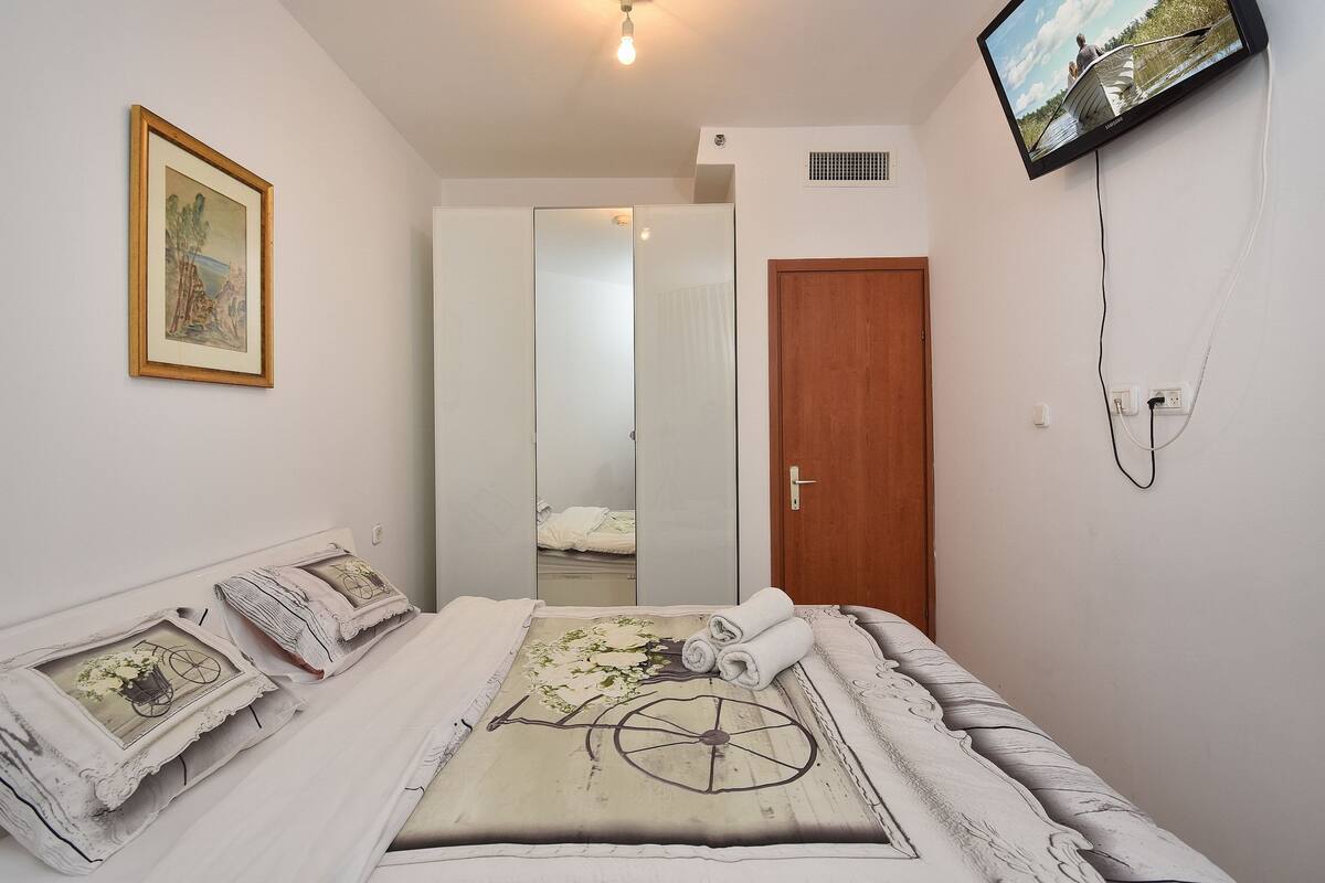 A bedroom with a bed, a TV mounted on the wall, a wooden door, and a framed picture on the wall.
