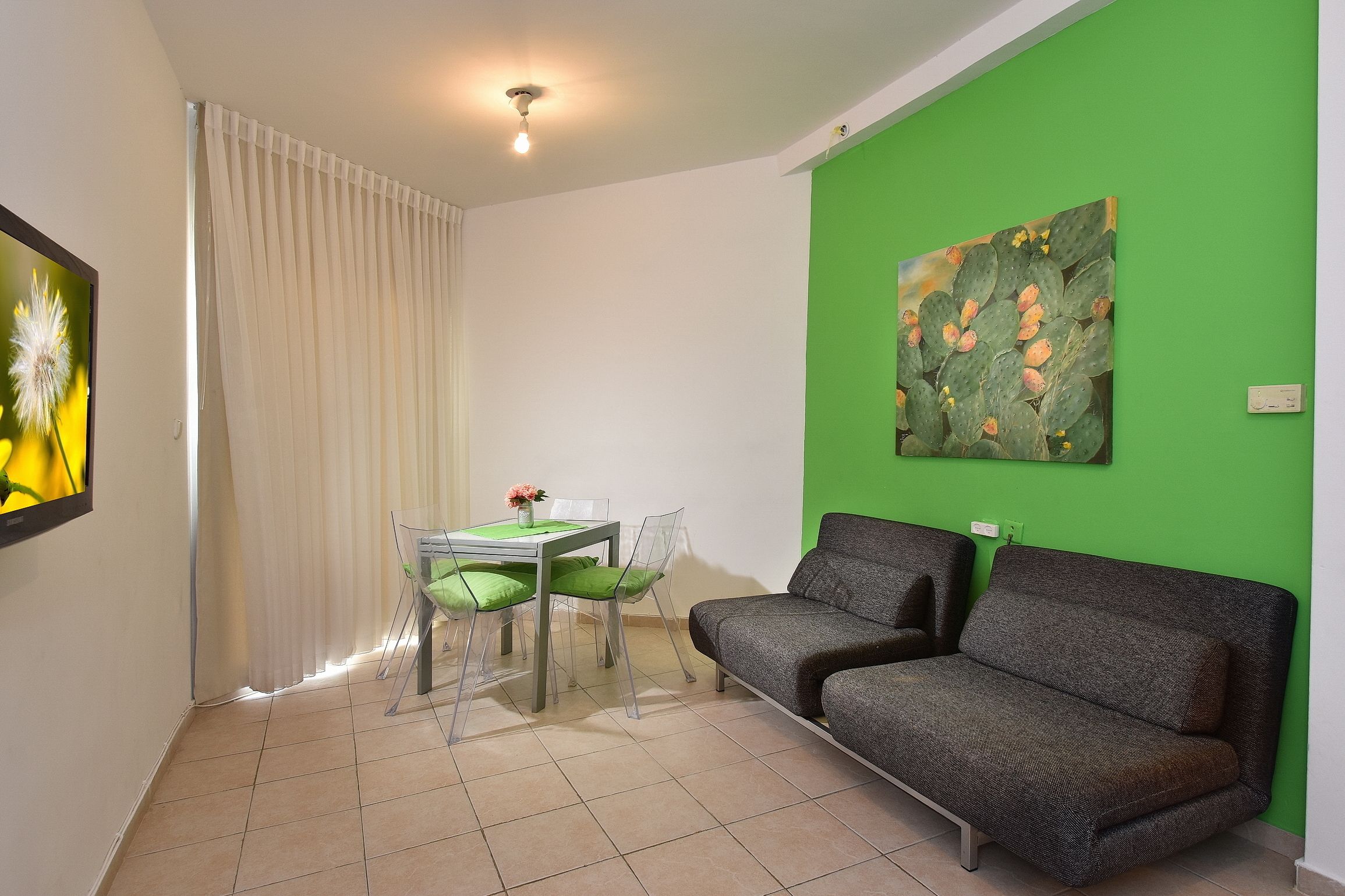 A living room with a green wall, a grey sofa, a dining table with chairs, and a painting of cacti.