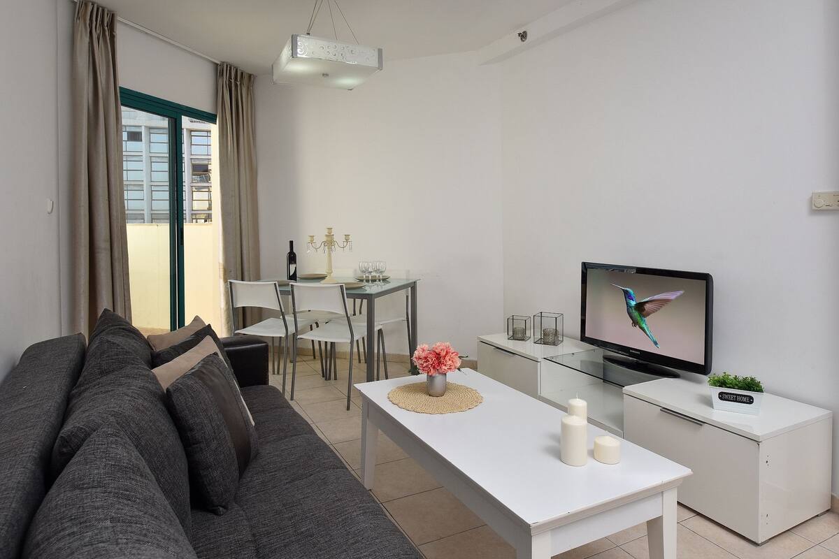 A modern living room with a grey sofa, a white table, a TV, and a dining area with a table and chairs.