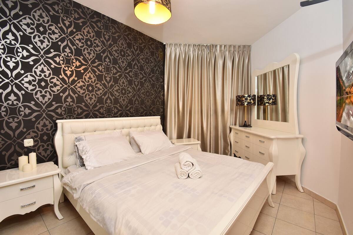 A neatly made bed with a white headboard, a bedside table with a lamp, and a wall with a patterned wallpaper.