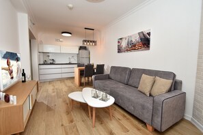 Ein modernes Wohnzimmer mit grauem Sofa, einem weißen Couchtisch und einem Holznachttisch. Im Hintergrund befindet sich eine Küche mit weißen Schränken und einem Essbereich.