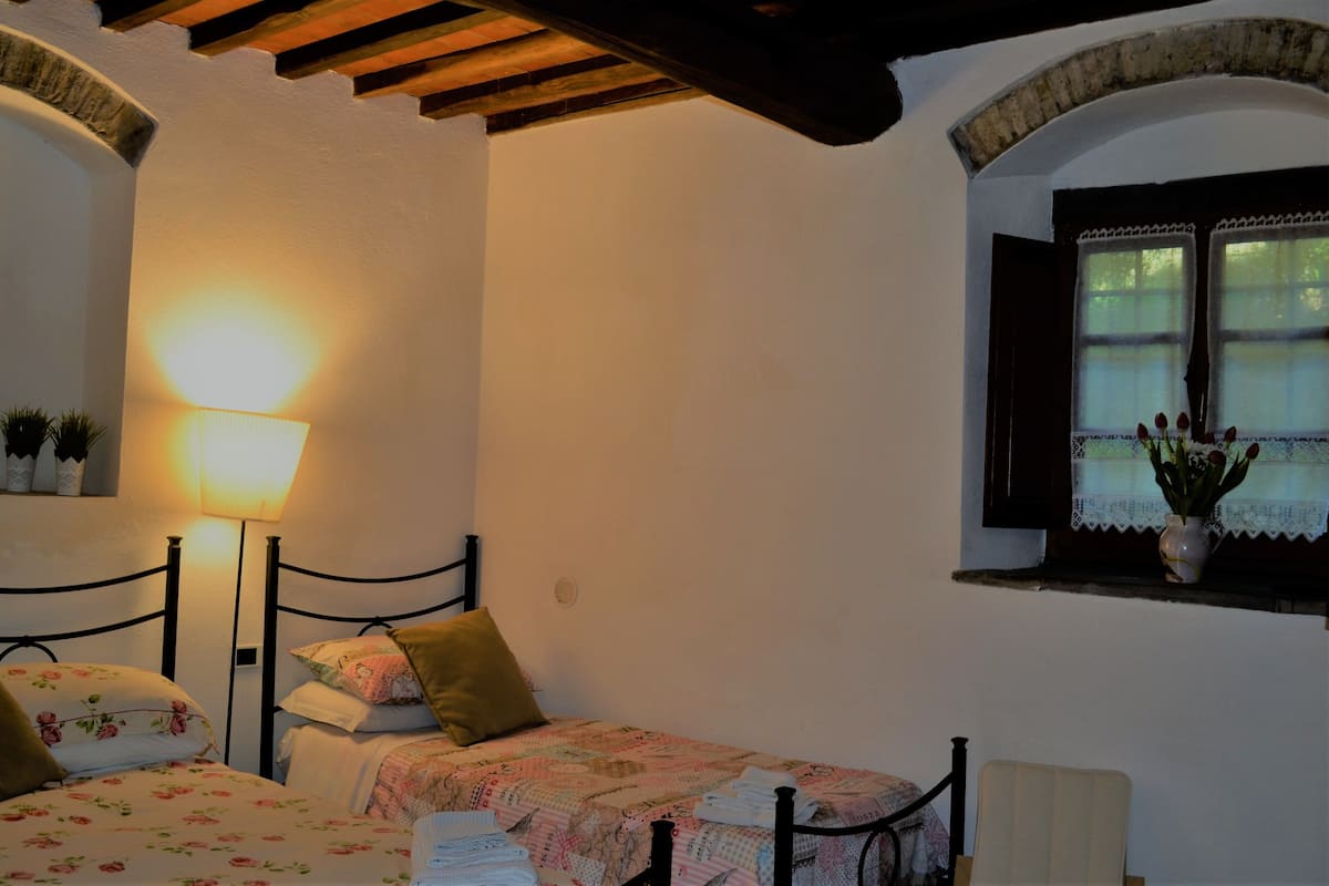 Ein Schlafzimmer mit zwei Betten, einer Lampe, einem Fenster mit Blumen und einem kleinen Regal mit Pflanzen.