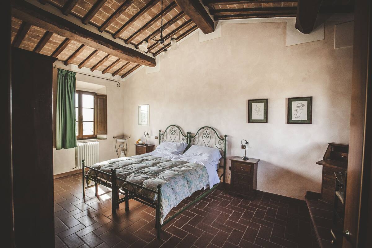 Ein Schlafzimmer mit einem Holzbettgestell, einem Heizkörper, einem Fenster mit grünen Vorhängen und zwei gerahmten Bildern an der Wand.