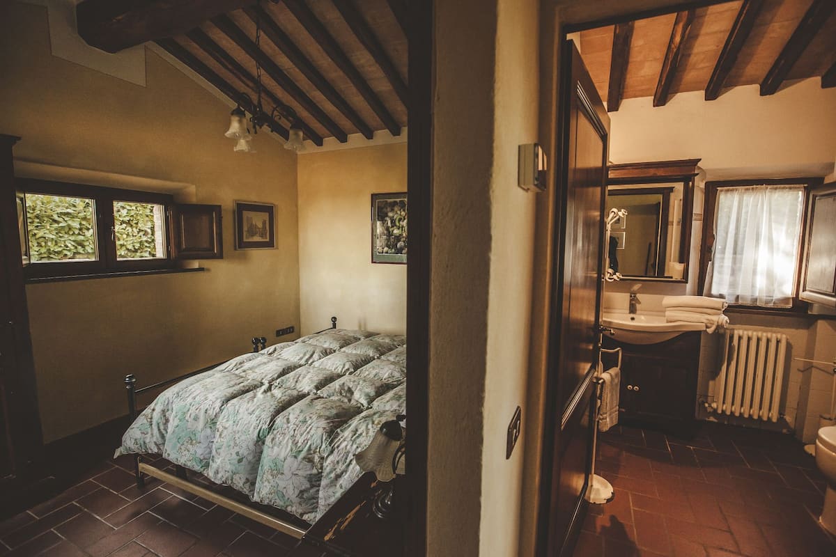 Ein Schlafzimmer mit Bett, Fenster und einem Badezimmer, das ein Waschbecken und einen Spiegel beinhaltet.