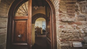 An arched wooden door leading into a room with a bed and a dresser. A stone wall with a plaque is visible to the right. - Zampugna (Trequanda)