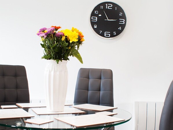 Un salón de conferencias con una mesa redonda de cristal, dos sillas tapizadas en gris, un florero blanco con flores y un reloj de pared negro.