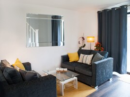 Uma sala de estar moderna com um sofá cinza, uma mesa de centro de vidro e um espelho grande na parede.
