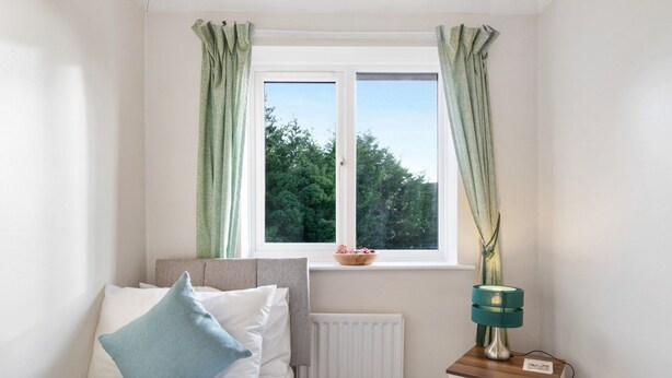 Une chambre à coucher comprenant un lit, une table de chevet, une lampe, une fenêtre avec des rideaux et une vue sur les arbres à l’extérieur.