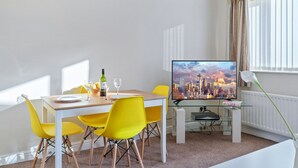 Un espace de repas moderne avec une table en bois, des chaises jaunes, une bouteille de vin et un écran de télévision diffusant des images de la ville.
