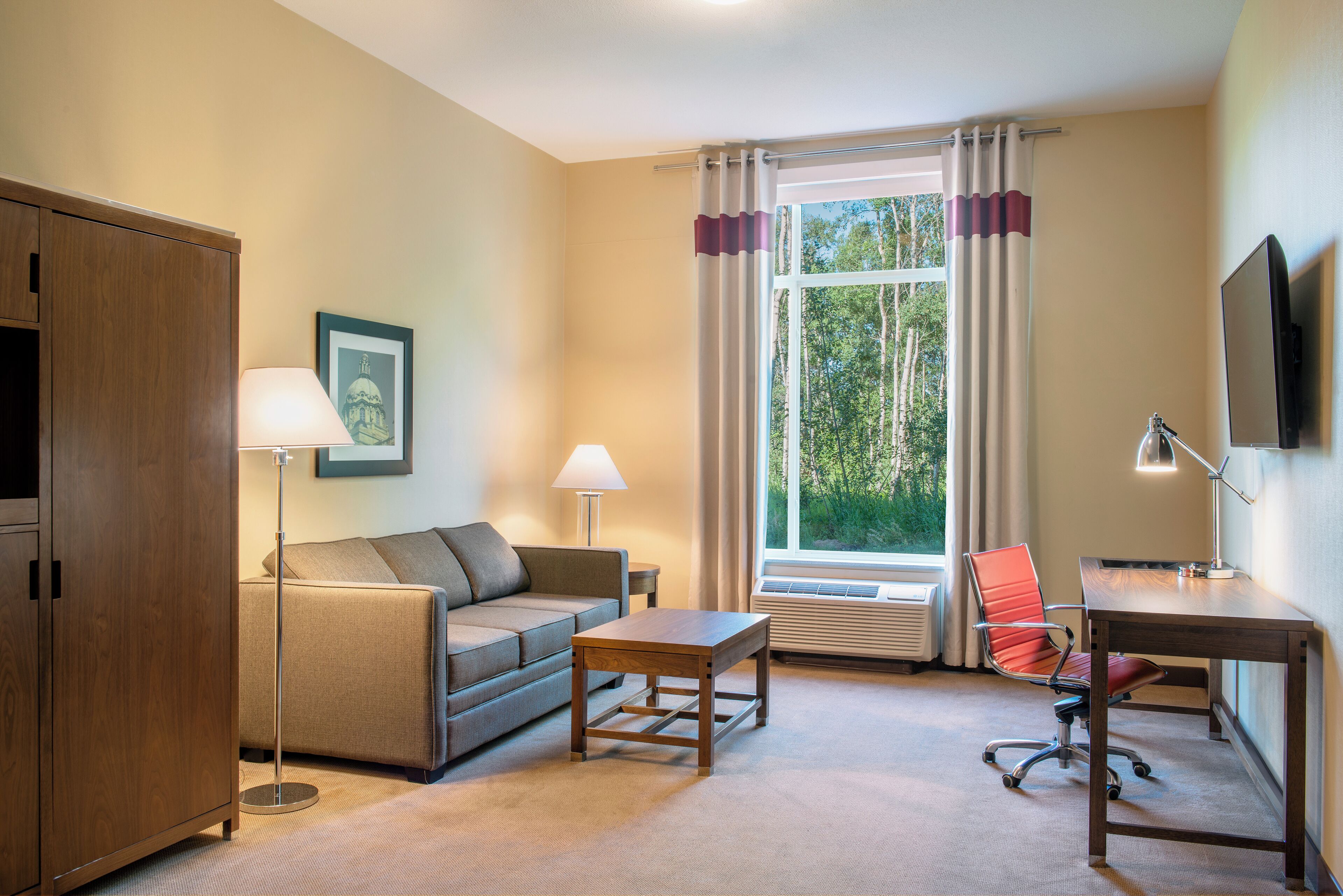 A furnished room with a sofa, a desk, a chair, a lamp, a picture on the wall, and a window with curtains.