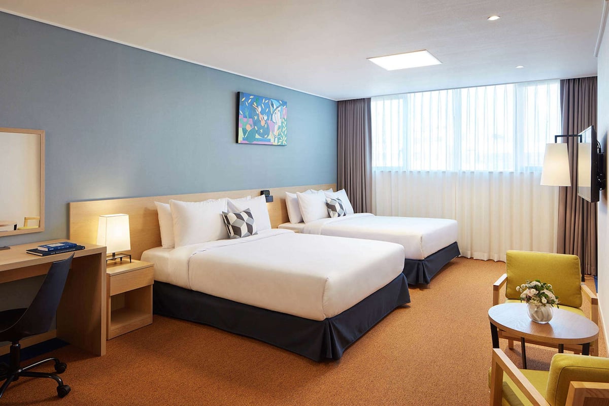 A hotel room with two beds, a desk, a chair, a small table with a vase, and a painting on the wall.