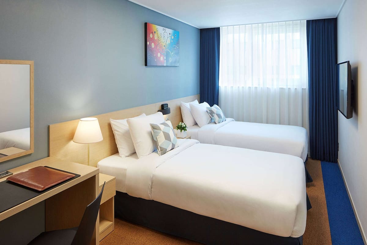 A hotel room with two beds, a desk, a chair, a lamp, a TV, and a painting on the wall.