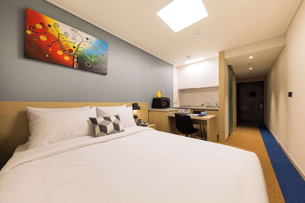 A hotel room with a bed, a desk, a microwave, and a painting on the wall.