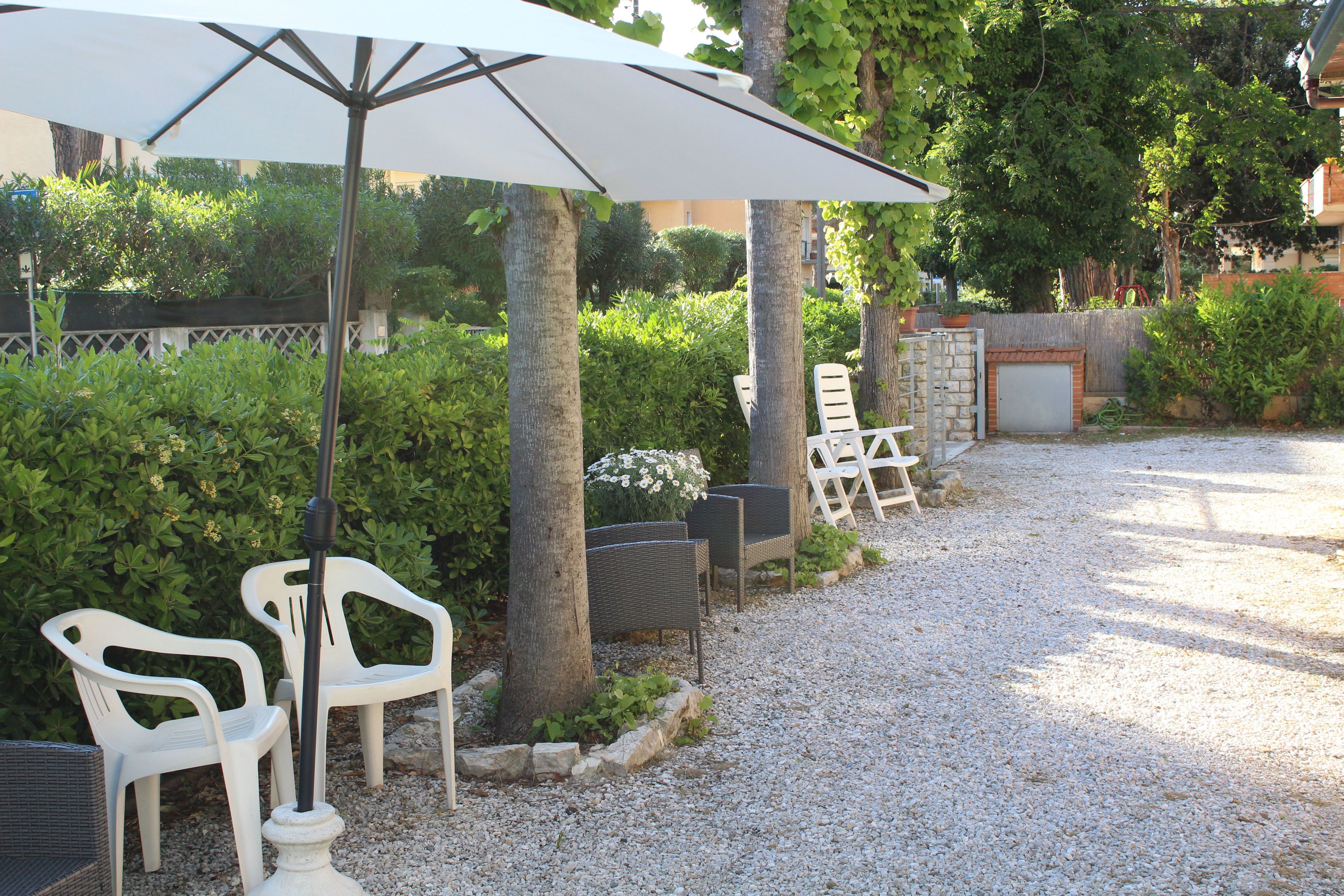 Eine Terrasse mit weißen Kunststoffstühlen, einem großen Sonnenschirm und einem Kiesweg.