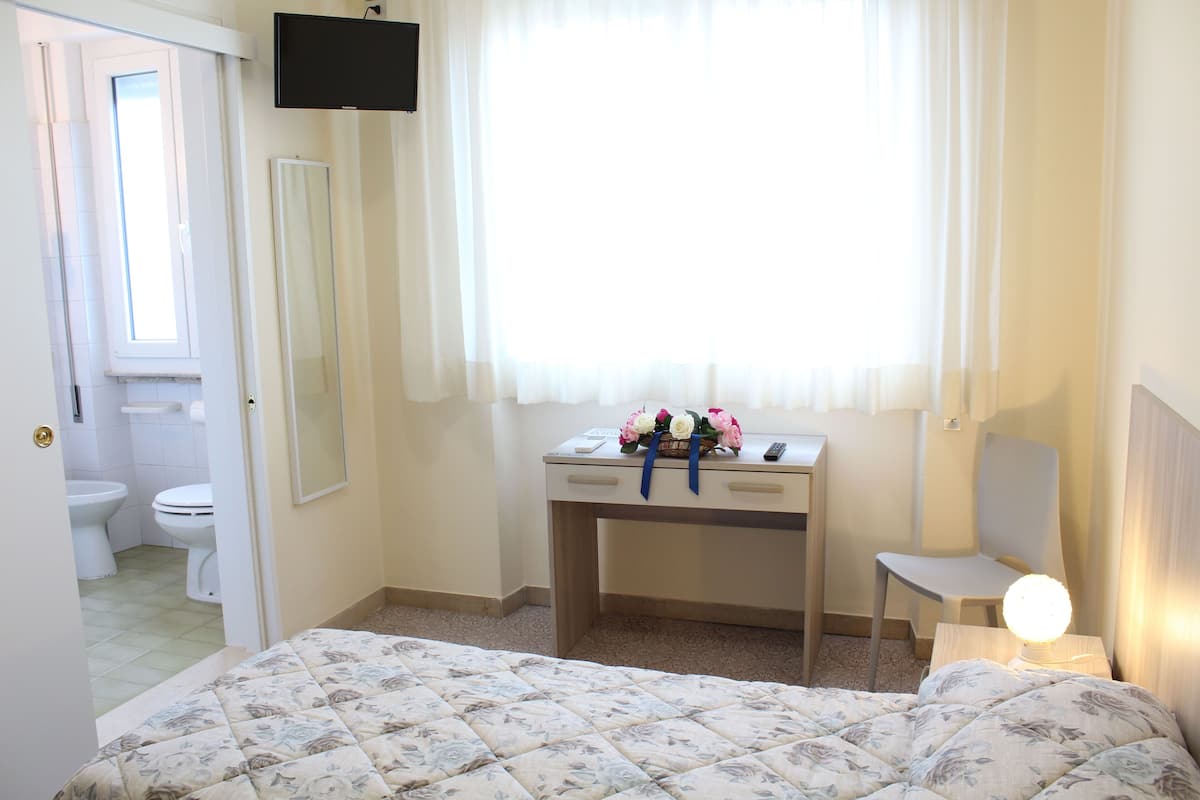 A bedroom with a bed, a desk with a lamp, a chair, a window with sheer curtains, and a bathroom with a toilet and sink.