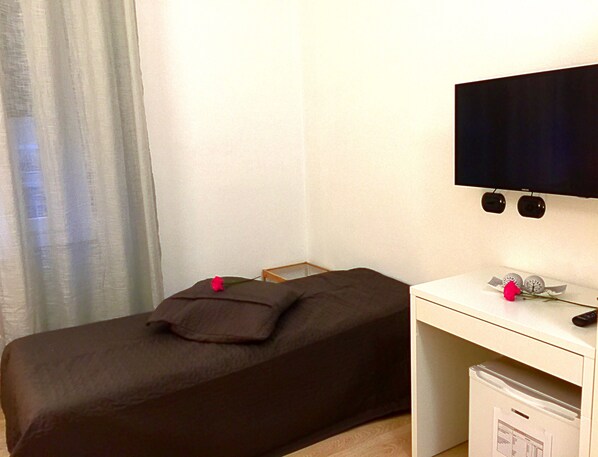 Una habitación pequeña y ordenada con una cama individual, un escritorio con televisor y un refrigerador pequeño.