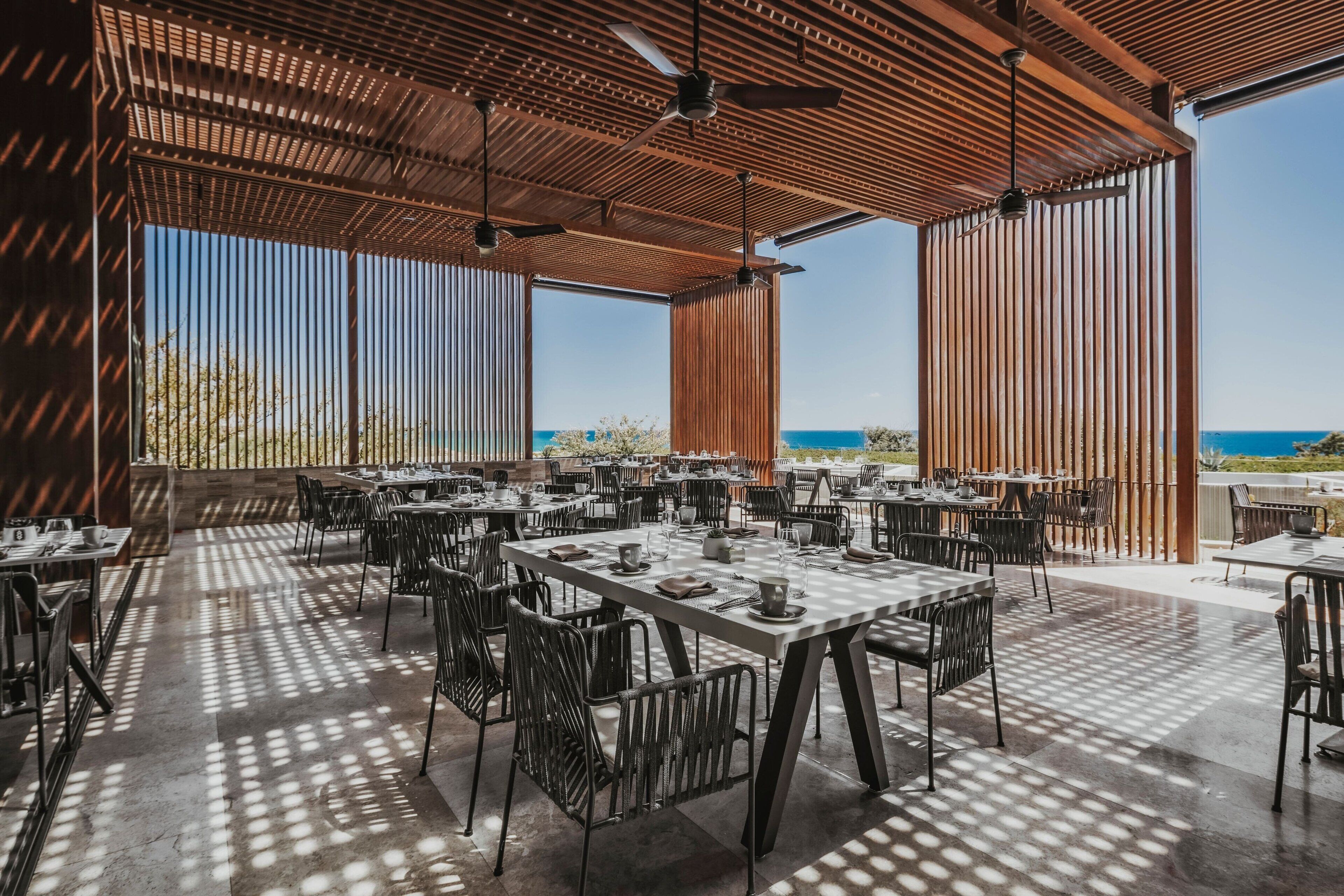 An outdoor dining area with tables and chairs, large windows, and a view of the sea.