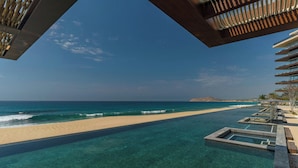 A clear view of a beach with a pool extending from the shore, a wooden deck, and a modern building with balconies. - Solaz, A Luxury Collection Resort, Los Cabos a Marriott Hotel (San José del Cabo)