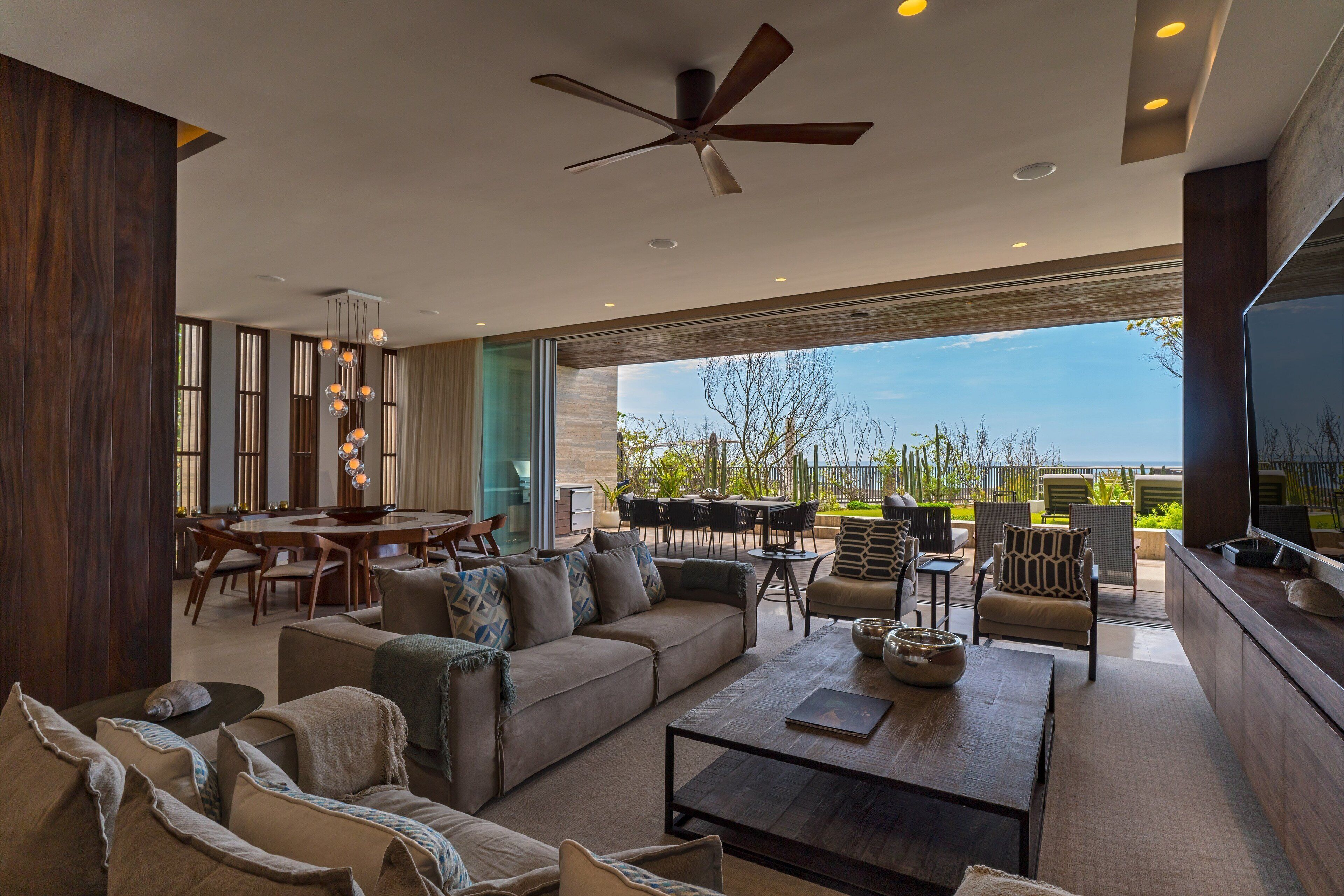 A spacious living area with a large sofa, coffee table, and dining set. There is a ceiling fan, large windows, and a view of the outdoors.