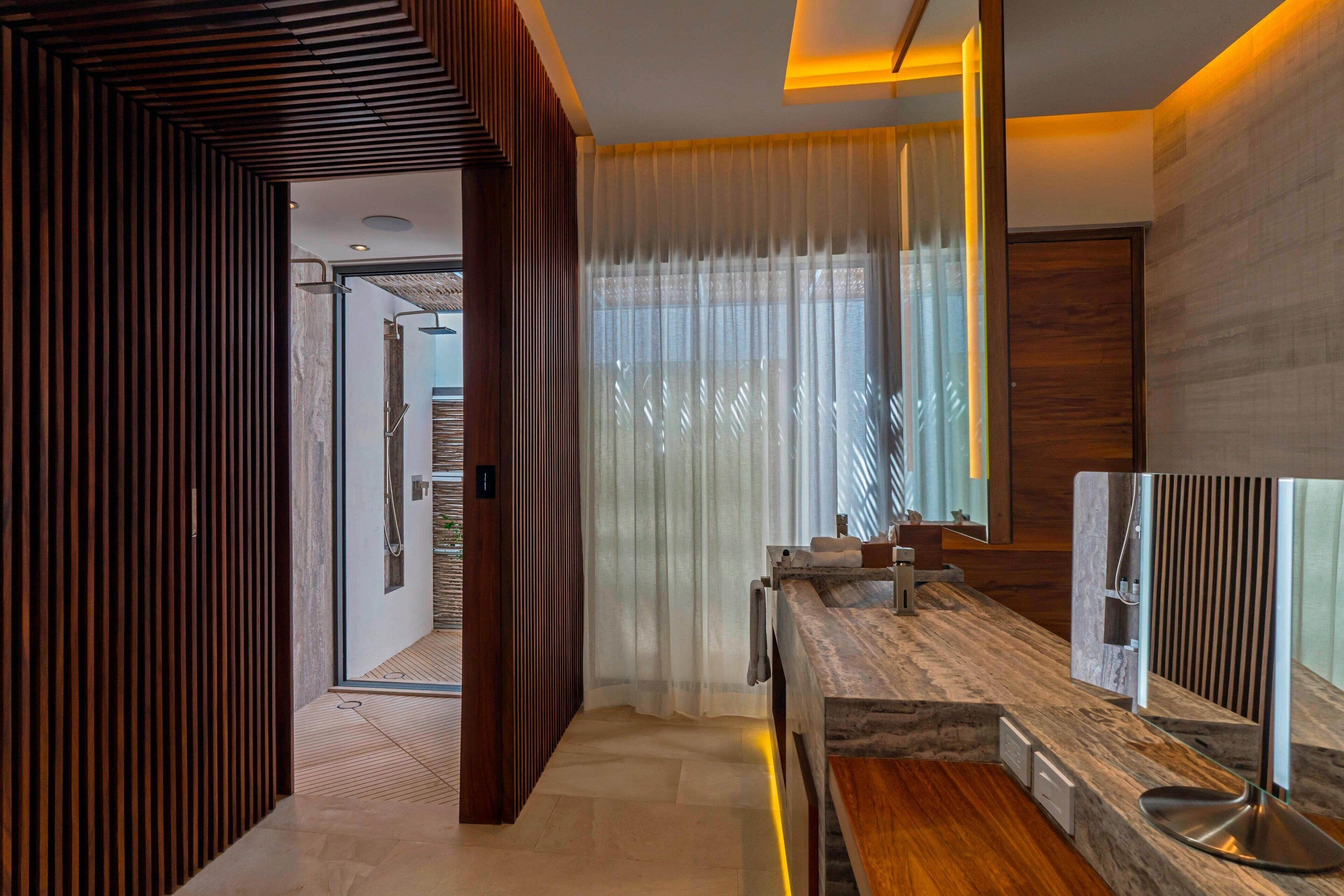 Un baño moderno con tocador de madera, un espejo grande y una zona de ducha visible a través de una puerta de vidrio.