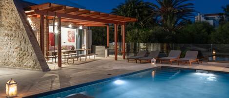 A pool area with lounge chairs, a pergola, and a stone wall.