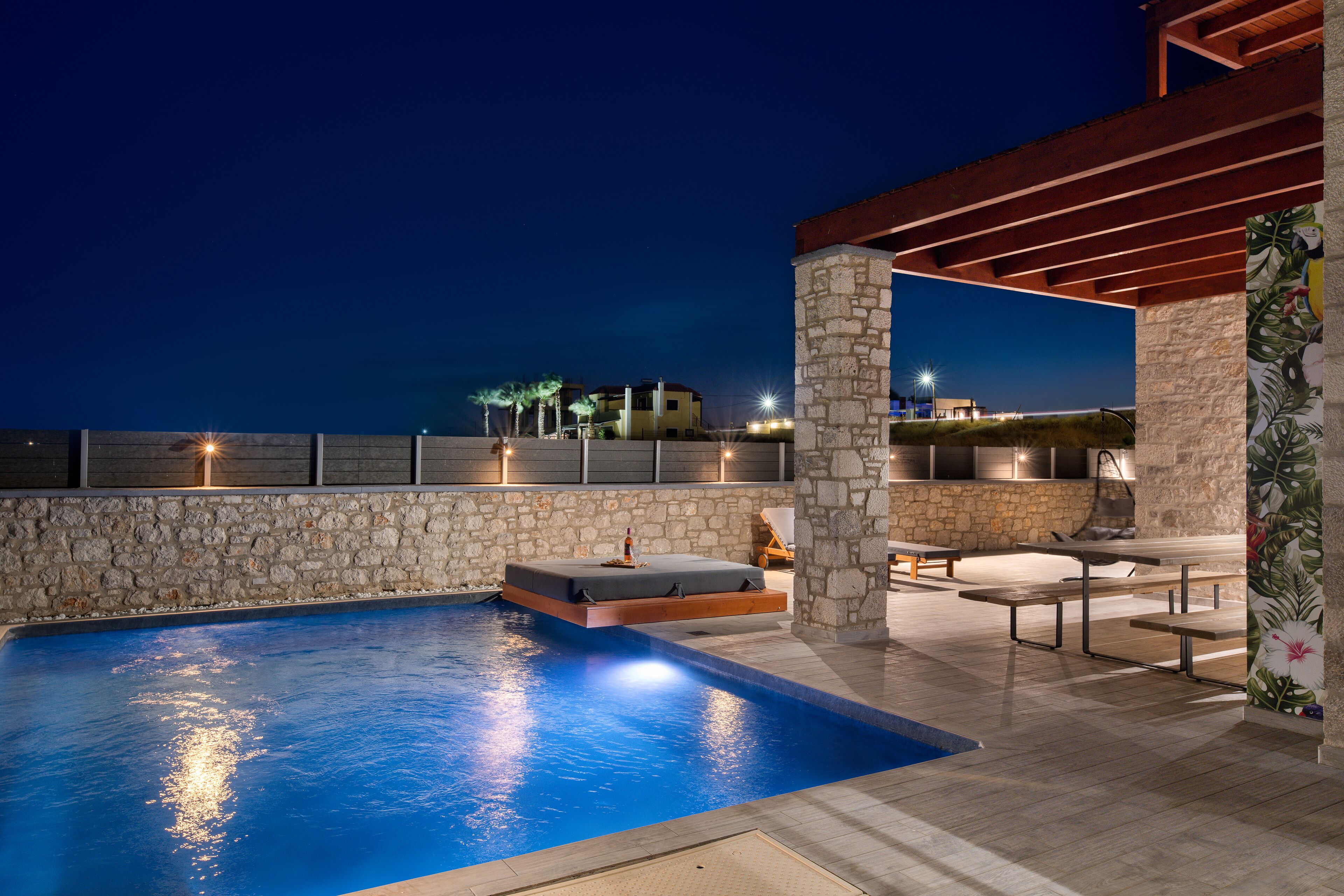 A swimming pool with a stone wall and wooden deck, benches, and a view of the sea at dusk.