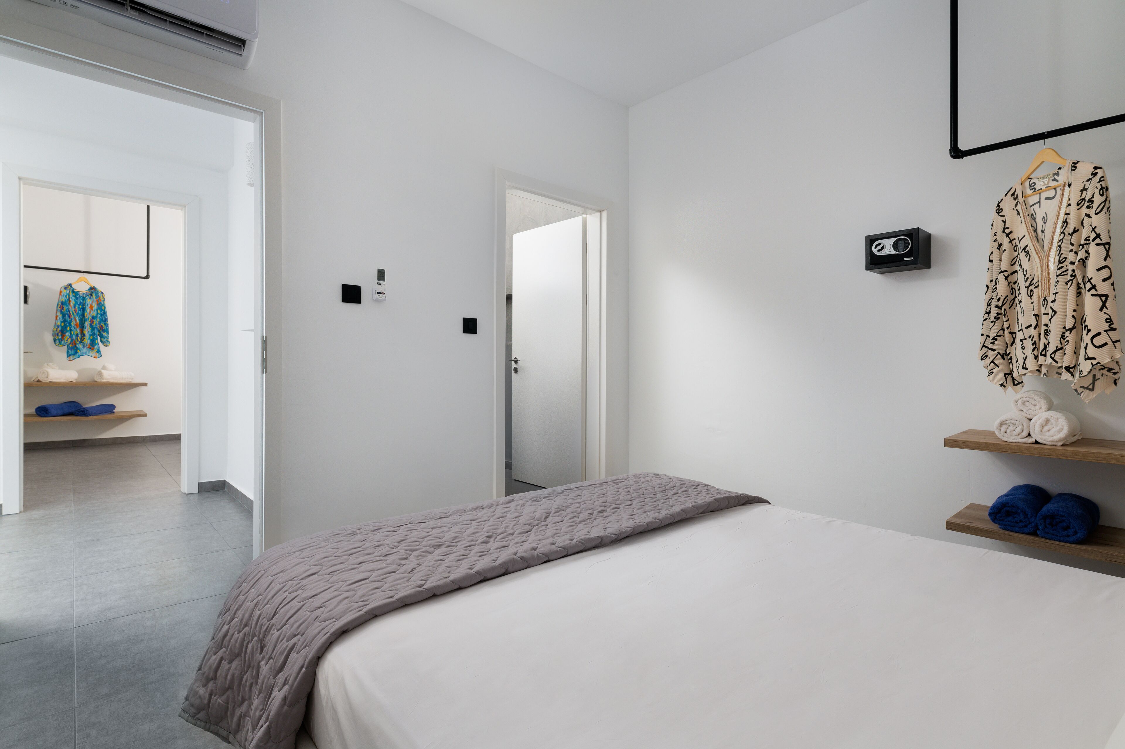 A modern hotel room with a bed, a bedside table, a wall-mounted radio, and a wardrobe with hanging clothes.