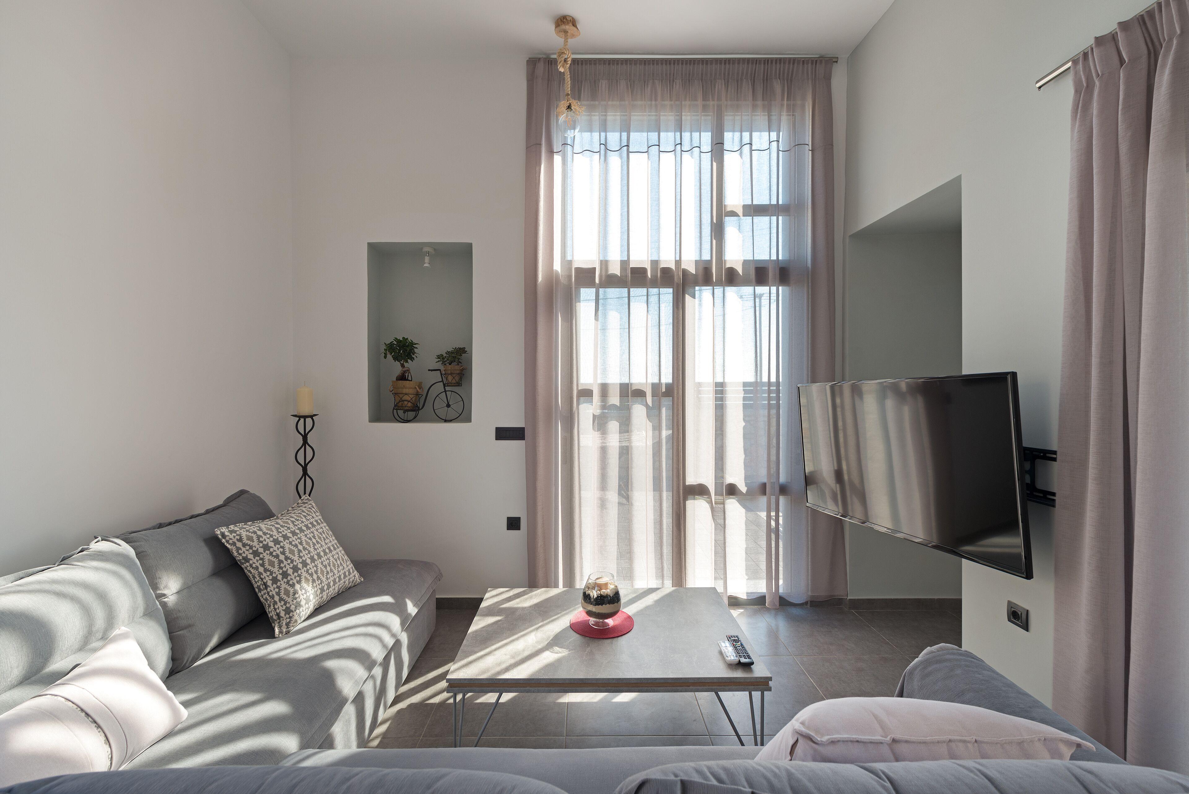 Un salon moderne comprenant un canapé, une table basse agrémentée d’un objet décoratif, un téléviseur à écran plat fixé au mur et une grande fenêtre avec des rideaux.