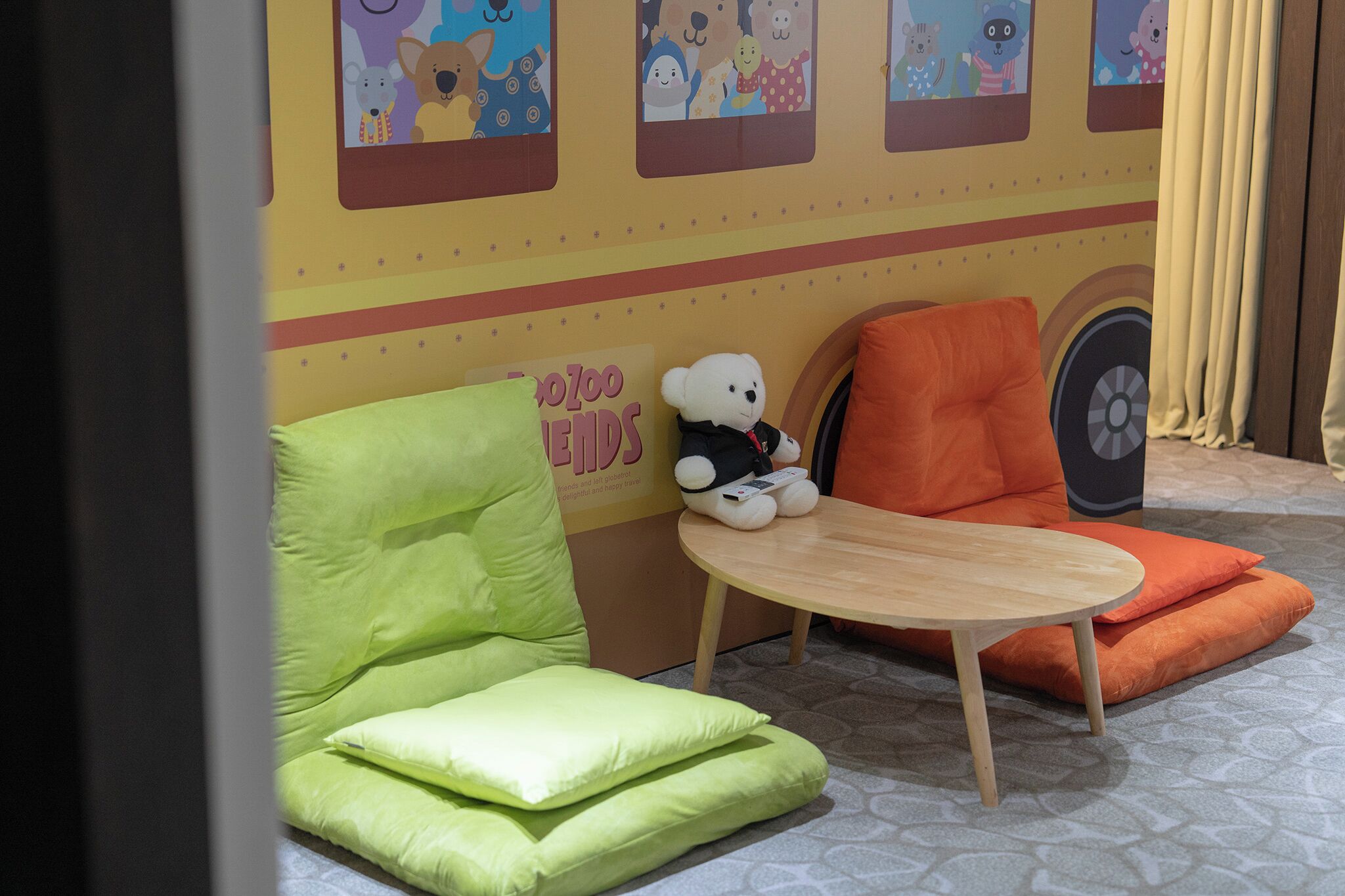 Zona infantil con asientos acolchados verde y naranja, una mesita y una pared decorada con personajes de dibujos animados.