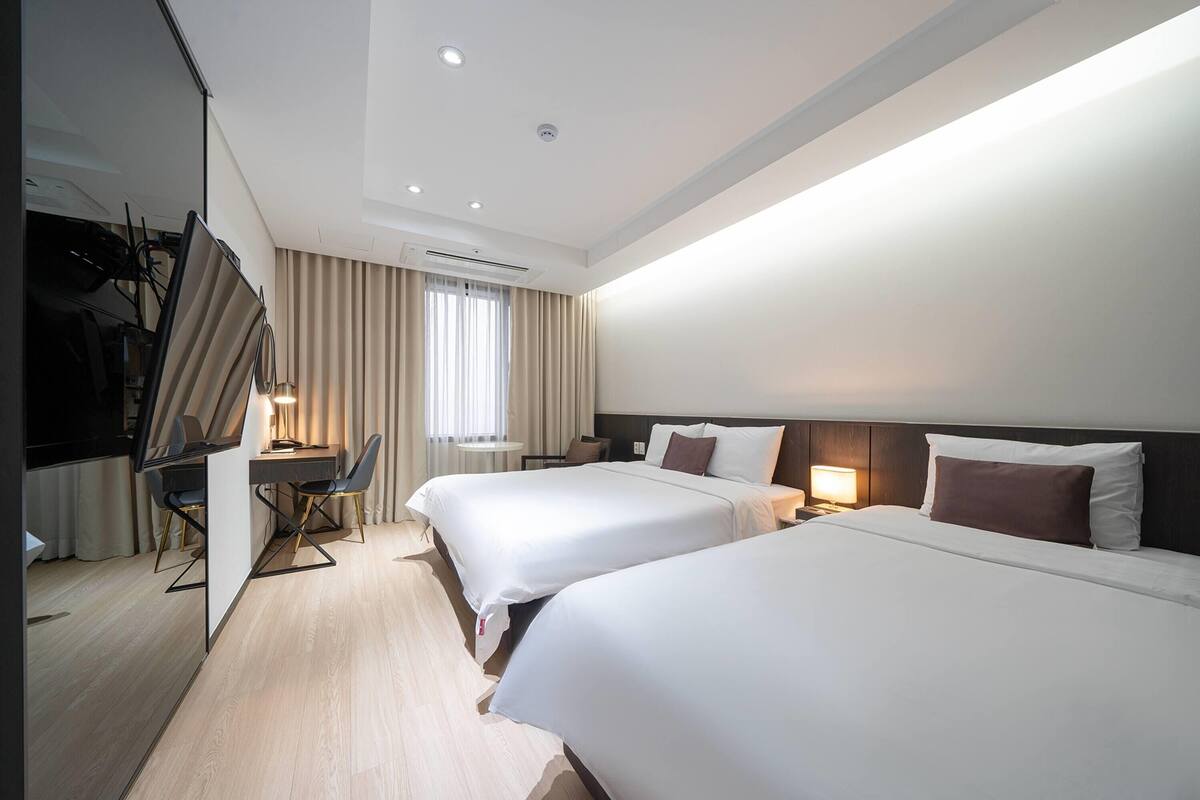 A hotel room with two beds, a desk, a TV, and a window with curtains.