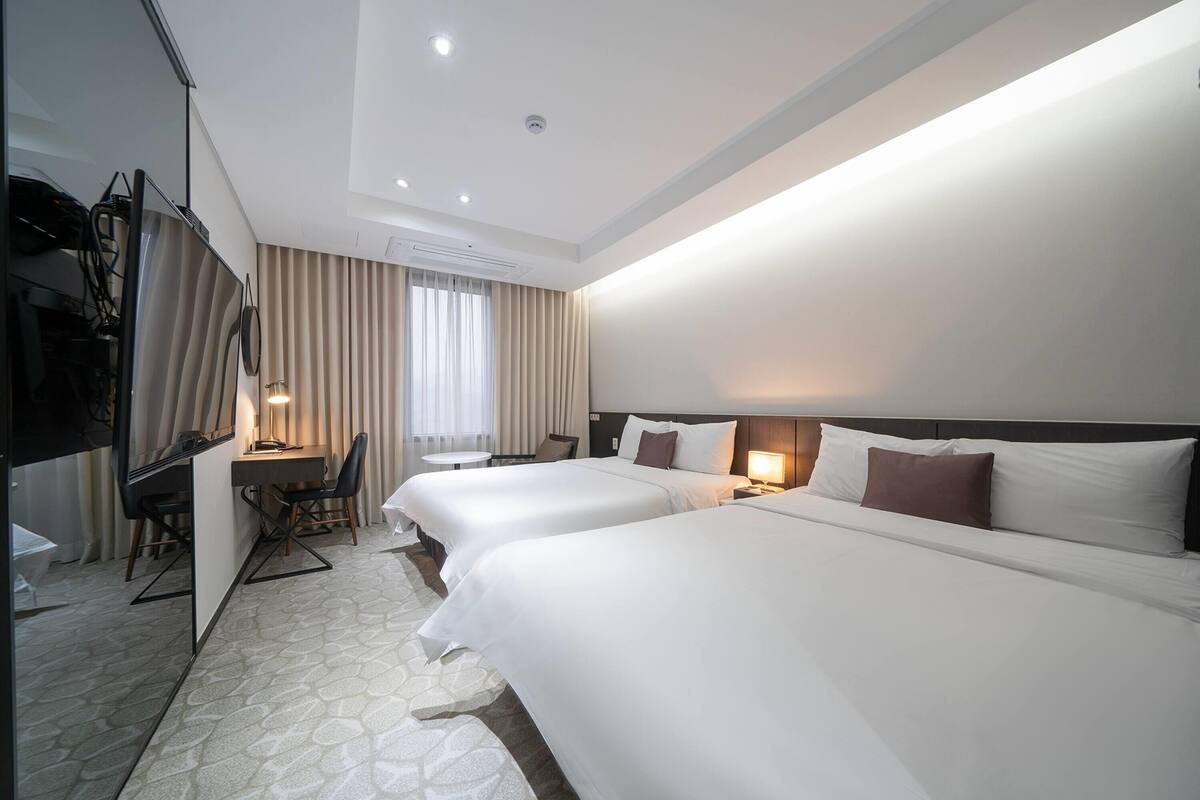 A hotel room with two beds, a desk, a chair, a TV, and a window with curtains.