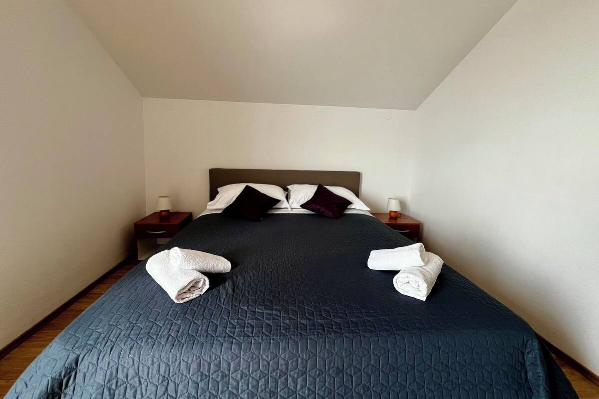 A neatly made bed with a dark quilt, two pillows, and two rolled towels on the bed.